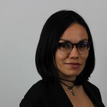 A headshot of a woman with black hair, glasses, and a calm expression on a grey background.